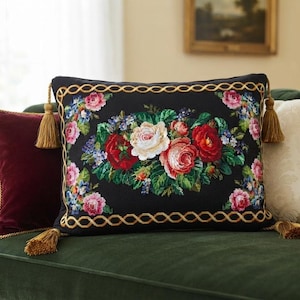 May include: A rectangular black throw pillow featuring a central floral arrangement of red, white, and pink roses with green leaves. The pillow is bordered by a gold rope pattern and finished with gold tassels at each of the four corners.