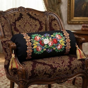 May include: A black cylindrical bolster pillow featuring a central embroidered floral bouquet of red, pink, and white roses with yellow accents, flanked by gold patterned borders and finished with decorative gold and red tassels on each end.