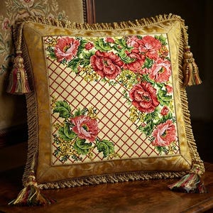 May include: A square decorative pillow featuring a central panel with a red lattice pattern and embroidered pink floral clusters. The pillow is framed by a gold border and finished with gold twisted cord trim and tassels at each corner.