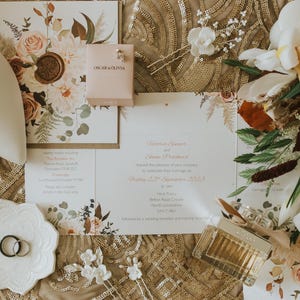 Pu&ograve; includere: Un set di inviti di nozze con un design floreale, che comprende un bouquet di fiori arancioni e bianchi, un paio di scarpe bianche e una piccola scatola con il testo "Oscar & Olivia".