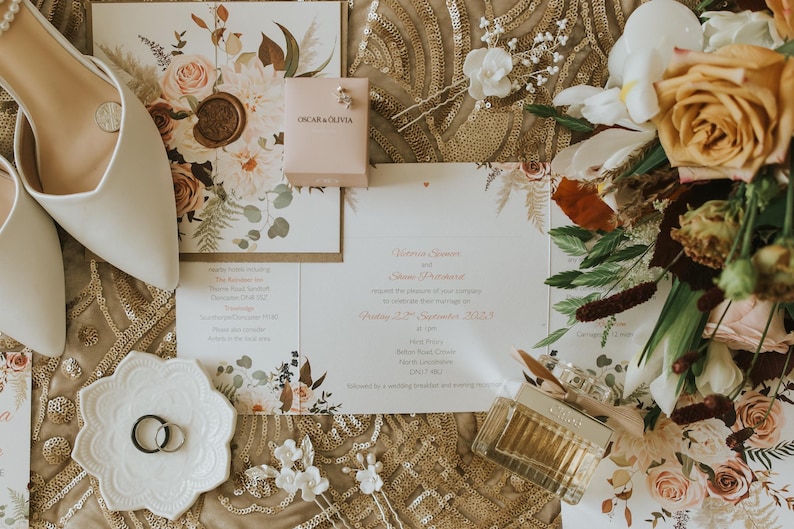 Pu&ograve; includere: Un set di inviti di nozze con un design floreale, che comprende un bouquet di fiori arancioni e bianchi, un paio di scarpe bianche e una piccola scatola con il testo "Oscar & Olivia".