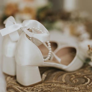 Pu&ograve; includere: Scarpe da sposa in raso bianco con cinturino alla caviglia di perle e un fiocco sul davanti. Le scarpe sono su uno sfondo di tessuto marrone.