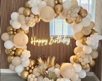 Pode incluir: Um arco de balões circular em tons de branco, dourado e bege, com uma placa de néon "Happy Birthday". O arranjo inclui balões de vários tamanhos e elementos decorativos, criando um cenário festivo.