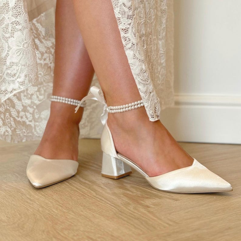 Pu&ograve; includere: Scarpe da sposa in raso bianco con punta a punta e tacco a blocco. Le scarpe hanno un cinturino alla caviglia con perle e un fiocco di raso.
