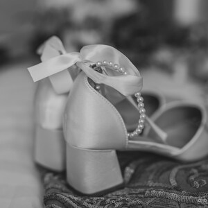 Pu&ograve; includere: Un paio di scarpe da sposa in raso bianco con un ornamento di perle e nastro. Le scarpe sono su un tessuto a fantasia.