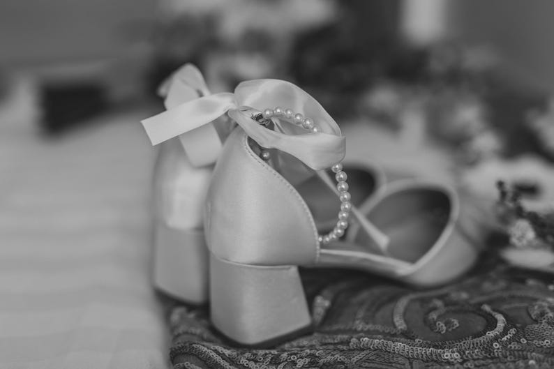 Pu&ograve; includere: Un paio di scarpe da sposa in raso bianco con un ornamento di perle e nastro. Le scarpe sono su un tessuto a fantasia.