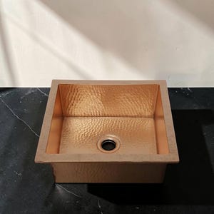 Op de afbeelding: Een gehamerde koperen keukenwasbak met een vierkant bassin en een centrale afvoer. De wasbak heeft een roodbruine kleur en een getextureerd oppervlak. Ontworpen voor keukengebruik, meet het ongeveer 40 cm x 40 cm.