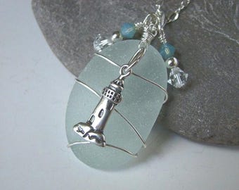 May include: A silver lighthouse charm hangs from a light blue sea glass pendant wrapped in silver wire. The pendant is attached to a silver chain with blue and clear glass beads.