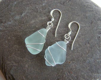May include: A pair of dangle earrings featuring smooth, frosted pale seafoam green glass pieces. Each piece is wrapped in thin, polished silver-colored wire and attached to a simple silver-colored metal hook for pierced ears.