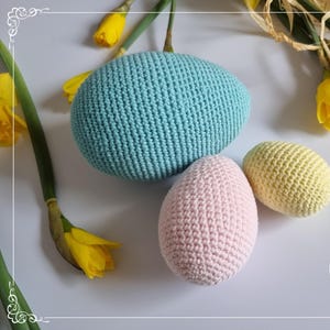 May include: Three crocheted Easter eggs in pastel colours: light blue, pink and yellow. The eggs are sitting on a white surface.
