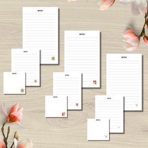 May include: White lined notepads and sticky notes with the word "NOTES" printed at the top. The notepads have gold snowflake, red and brown geometric, and deer head designs. The notepads are arranged on a light wood-grain surface.