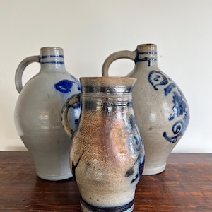 Op de afbeelding: Drie antieke steengoed kruiken met handvatten. De kruiken zijn in tinten grijs, bruin en blauw. De kruiken staan op een houten oppervlak. De kruiken hebben een rustieke, handgemaakte uitstraling.