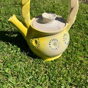 Può includere: Una teiera in ceramica gialla fatta a mano con un manico intrecciato e un coperchio abbinato. La teiera presenta disegni floreali in rilievo e un beccuccio. La teiera è appoggiata su un letto di erba verde.
