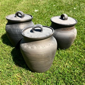 May include: Three handmade, dark gray ceramic pots with matching lids. Each pot has a rounded body and a loop handle on the lid. The pots are sitting on green grass in natural sunlight. The pots have a rustic, textured finish.