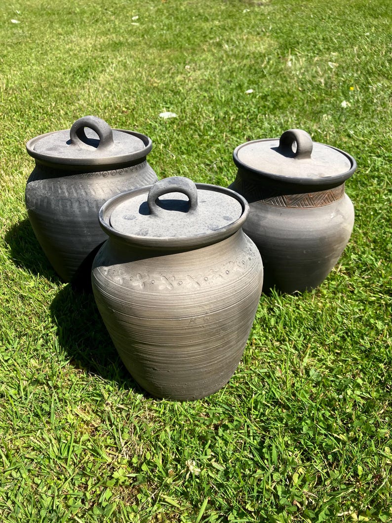 May include: Three handmade, dark gray ceramic pots with matching lids. Each pot has a rounded body and a loop handle on the lid. The pots are sitting on green grass in natural sunlight. The pots have a rustic, textured finish.