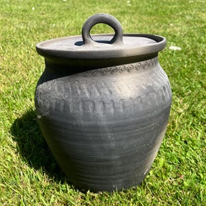 May include: A dark gray ceramic pot with a lid and a handle. The pot has a rounded shape with decorative patterns around the rim. The pot is sitting on green grass.