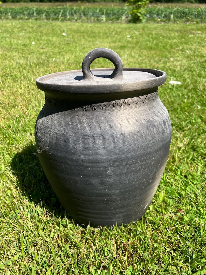 May include: A dark gray ceramic pot with a lid and a handle. The pot has a rounded shape with decorative patterns around the rim. The pot is sitting on green grass.