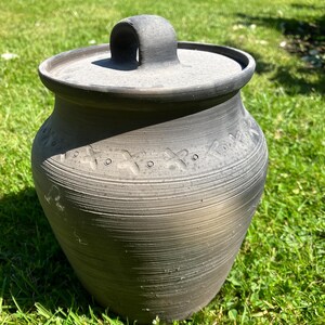 May include: A dark gray ceramic pot with a lid and handle, sitting on green grass. The pot has a textured surface with horizontal lines and decorative cross-shaped patterns near the top. The lid has a curved handle.