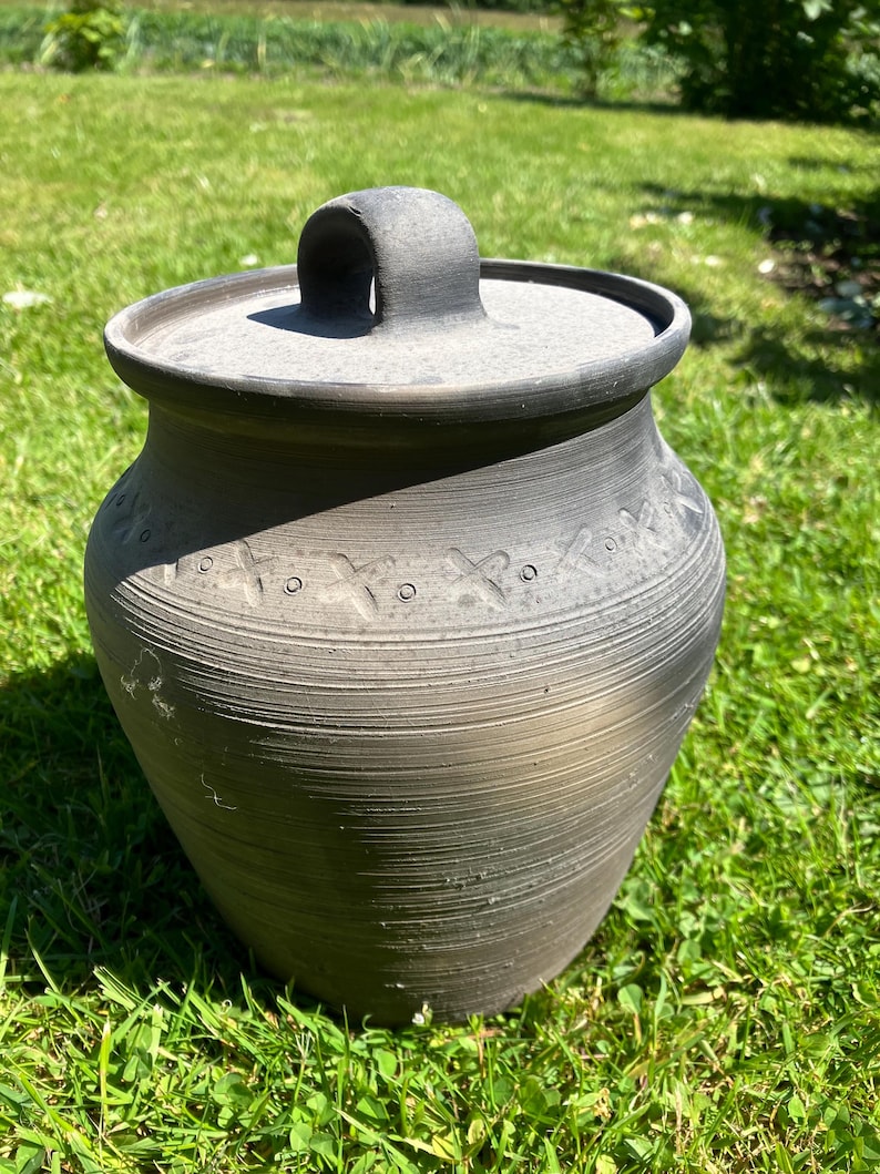 May include: A dark gray ceramic pot with a lid and handle, sitting on green grass. The pot has a textured surface with horizontal lines and decorative cross-shaped patterns near the top. The lid has a curved handle.