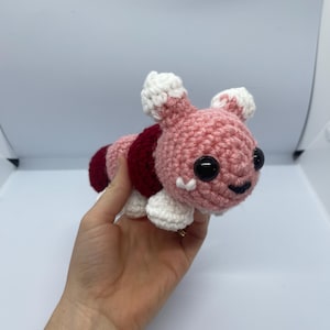 May include: A crocheted pink and red plush toy with white accents. The toy has a smiling face with black eyes and a small mouth.