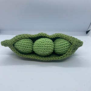 以下が含まれることがあります： 3つの緑色のエンドウ豆が入った、緑色の毛糸で編まれたエンドウ豆のさや