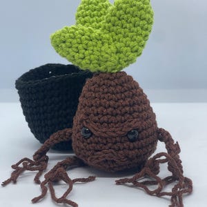 May include: A handmade crochet doll resembling a Mandrake plant, featuring a brown body, green leaves, and black eyes. The doll has brown root-like appendages and sits next to a black crochet pot. The background is plain white.
