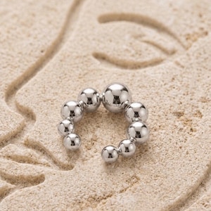 Könnte beinhalten: Ein silberfarbener Ohrschmuck mit einer Reihe von abgestuften, kugelförmigen Perlen. Der Schmuck wird auf einer strukturierten, hellbraunen Oberfläche mit einem dezenten, geschnitzten Design präsentiert. Die polierte Oberfläche des Ohrschmucks reflektiert das Umgebungslicht.