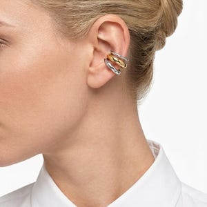 Puede incluir: Primer plano de una oreja adornada con un ear cuff triple. El ear cuff presenta tres bandas apiladas: plateada, dorada y plateada. La persona lleva una camisa blanca con cuello.