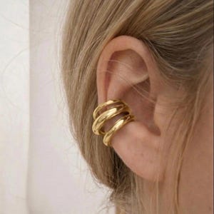 May include: A gold ear cuff with a layered, spiral design. The ear cuff wraps around the ear cartilage, creating a modern and stylish look. The jewelry is made of a shiny, metallic material.