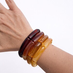 Op de afbeelding: Een stapel van vier dikke, doorschijnende armbanden in tinten amber, bruin en geel. De armbanden worden gedragen om een pols tegen een witte achtergrond. De armbanden zijn gemaakt van een glad, glanzend materiaal.