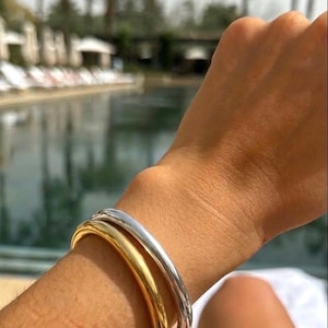 Può includere: Primo piano di un polso adornato da tre braccialetti in metallo. Uno è dorato, gli altri due sono argentati. I braccialetti sono lisci e hanno un design arrotondato. Lo sfondo mostra una piscina e palme.