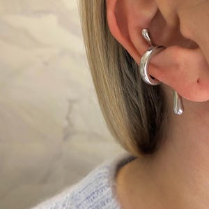 Peut inclure: Un ear cuff argenté avec un design unique et fluide. Le bijou entoure l'oreille et présente une forme de goutte d'eau pendante. Le bijou est en métal brillant.