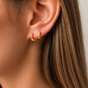 Può includere: Un paio di piccoli orecchini a cerchio dorati e lucidi con un profilo a bordi quadrati. Gli orecchini sono indossati sul lobo dell'orecchio e presentano una finitura metallica liscia e riflettente con un semplice design a cerniera.