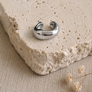 May include: A polished silver-tone ear cuff with a smooth, rounded, and thick C-shaped design. The metal surface is reflective and features a continuous, solid band that curves into an open loop, creating a simple and bold accessory for the ear.