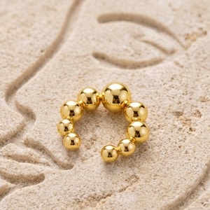 Peut inclure: Un ear cuff doré composé d'une série de perles sphériques graduées. Le bijou est présenté sur une surface texturée et claire, créant un contraste avec le métal brillant.
