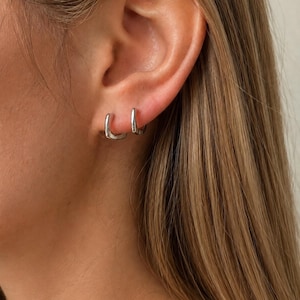 Peut inclure: Une paire de petites créoles argentées au profil carré. Ces boucles d'oreilles présentent une finition métallique lisse et réfléchissante, conçues pour se positionner près du lobe de l'oreille, offrant un aspect épuré et simple.
