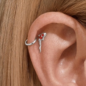May include: A silver ear cuff and a koi fish earring are shown on an ear. The koi fish earring features a red and white design. The ear cuff is a simple, curved silver band. The background is a person's skin and hair.