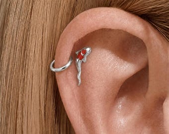 May include: A silver ear cuff and a koi fish earring are shown on an ear. The koi fish earring features a red and white design. The ear cuff is a simple, curved silver band. The background is a person's skin and hair.
