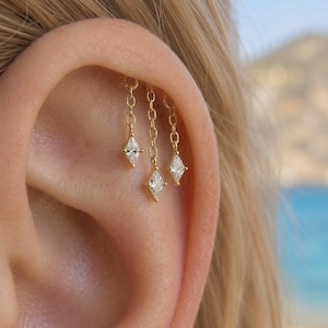 Peut inclure: Un bijou d'oreille en or composé de trois chaînes pendantes délicates, chacune terminée par une petite pierre précieuse transparente de taille marquise. Le bijou est fixé au cartilage supérieur de l'oreille, les chaînes pendant verticalement.