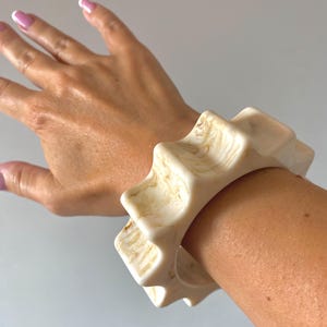 Può includere: Bracciale spesso color crema con un design geometrico a forma di stella. Il bracciale ha un effetto marmorizzato con venature marrone chiaro. Il bracciale è indossato al polso.