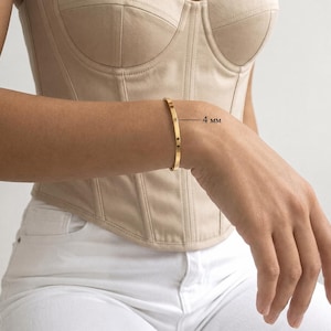 Può includere: Un bracciale rigido color oro, liscio e lucido, con un profilo piatto. Il bracciale presenta un design a fascia continua e pulita. Una linea sottile indica il lato del bracciale con il testo 4 mm, che indica la larghezza della fascia.