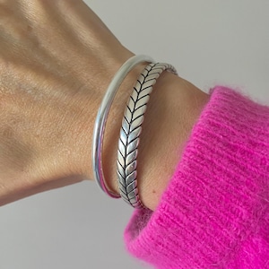 Puede incluir: Dos pulseras de plata apiladas en una muñeca. Una es una banda lisa y sencilla, y la otra presenta un diseño texturizado en espiga. La muñeca es parcialmente visible, y la manga de un suéter rosa brillante está en el fondo.