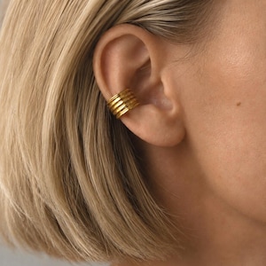 Puede incluir: Un ear cuff de tono dorado que presenta una serie de cinco crestas horizontales paralelas y apiladas. La pieza de joyería está colocada en el cartílago exterior de la oreja, mostrando un acabado metálico con una textura uniforme en la banda.