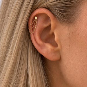 Op de afbeelding: Een goudkleurige kraakbeen-oorbel met een kleine ronde knop verbonden aan drie fijne, gedrapeerde kettinkjes. De kettinkjes hangen in een gelaagde formatie vanaf de knop, wat een gelaagd effect creëert langs de bovenrand van het oorkraakbeen.
