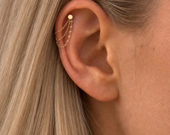 Può includere: Un orecchino da cartilagine color oro caratterizzato da un piccolo perno circolare collegato a tre catenelle sottili e drappeggiate. Le catenelle pendono in una formazione a strati dal perno, creando un effetto sovrapposto sul bordo superiore.
