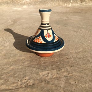 May include: A small ceramic tagine pot, a traditional Moroccan cooking vessel. It has a cream-coloured conical lid with blue and orange patterns, and a dark blue base with an orange bottom. The tagine is designed for slow cooking.
