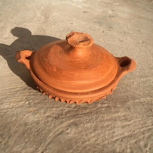 May include: A terracotta clay cooking pot with a lid. The pot has a round shape with a lid and two handles. The lid has a small knob on top. The pot is a warm orange-brown color and sits on a concrete surface.