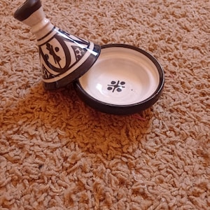 Può includere: Un tajine in ceramica, una tradizionale pentola da cucina marocchina, con coperchio conico e base poco profonda. Il coperchio e la base sono bianchi con motivi geometrici neri. Il tajine è appoggiato su una superficie marrone strutturata.