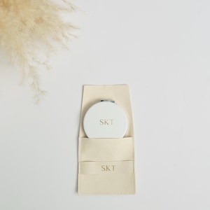 Peut inclure: Un miroir compact blanc avec une inscription dorée "SKT" est dans une pochette beige avec la même inscription.