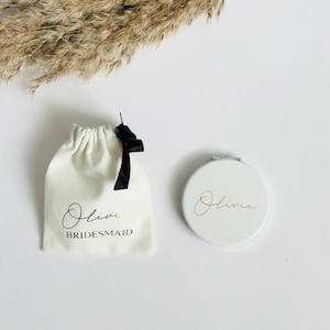 May include: A white fabric drawstring pouch with a black ribbon, featuring the gold script name Olivia and the black text BRIDESMAID, alongside a round white compact mirror with the gold script name Olivia printed on the front surface.